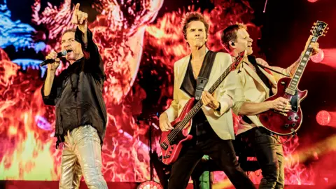Getty Images Three members of a band with a singer and two guitarists and a fire on a backdrop.