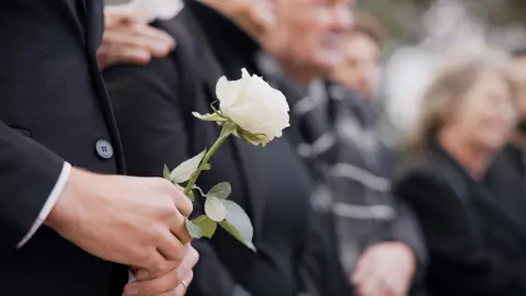 A group of people slightly out of focus. They are wearing black and attending a funeral. The person closest to the camera is holding a white rose. 