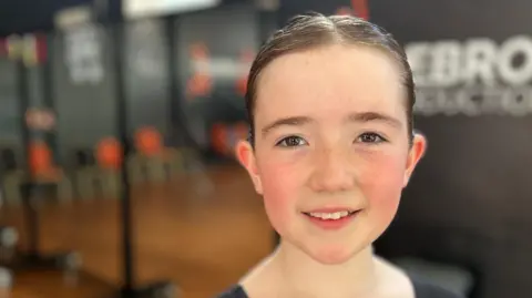 Ten year old Martha is looking at the camera, she is fair skinned with freckles and her hair is in a tight bun. Behind her is a dance studio. 