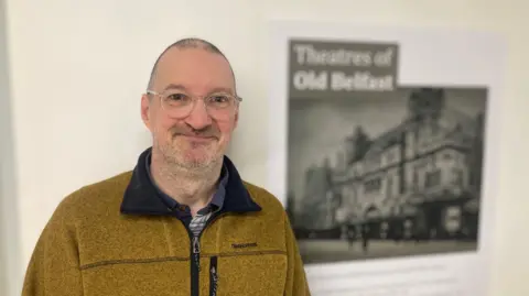 A man wearing round spectacles and a yellow fleece. He is smiling at the camera and standing in front of a poster which says 'Theatres of Old Belfast'. 