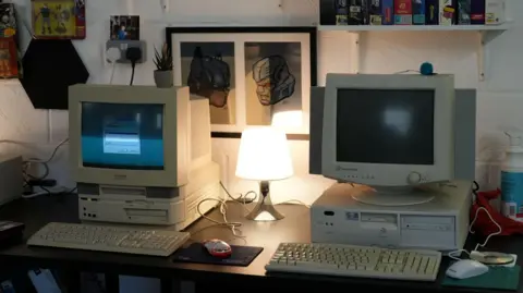 Shaun Whitmore/BBC Two retro computers side by side on a desk each with a mouse and keyboard. In between them is a lamp which is turned on.