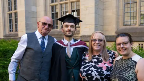 Paul Edwards dressed in graduation hat and grown, which has red drape around his suit. He is stood by his dad who is wearing suit trousers and a waistcoat and his mum who is wearing a white dress with black flowers. His girlfriend is wearing a white and black dress.
