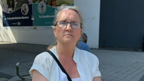 BBC/Jim Scott Sandra Goodsir, wearing black rimmed glasses and a white top, sits on a bench near the bus and Metro interchange in South Shields town centre. Behind her are two large posters. One on the left contains a directional arrow "To South Shields Market". On the right, a similar poster points "To Sandhaven". Both have images of people at the respective locations shown in photo-montages above the directional information.