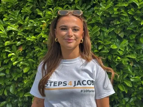 A young woman with long dark hair and gold hoop earrings smiles at the camera, while standing in front of a hedge. She is wearing a white t-shirt which says '#Steps4Jacob' and '#10forken' on it.