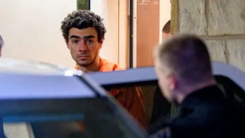 Man with brown curly hair in orange jumpsuit walks towards an open car door