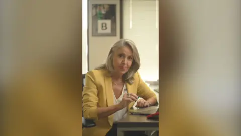 North America editor Sarah Smith wearing a yellow blazer. She's looking directly at the camera and raising her right hand.