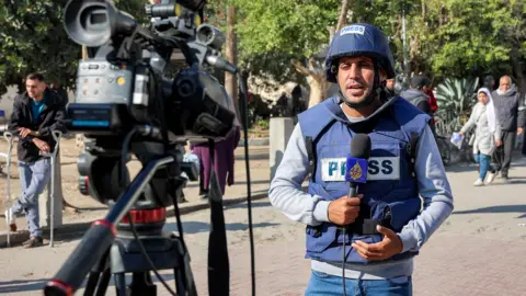 AFP Al Jazeera correspondent Mohammed Qreiqeh wears a press flak jacket and helmet while holding a microphone with the Al Jazeera logo on it, as he speaks towards a camera set up in the foreground of the image, in Gaza City in December 2024.