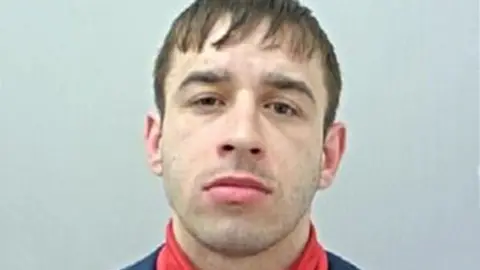 Police custody image of Terrence Kenny with brown hair and eyes wearing a red polo neck top and black jacket.