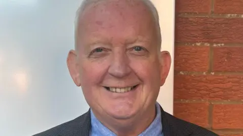 Reform A man with short white hair smiles at the camera. He is standing in front of a wall and wears a suit