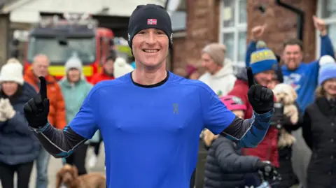 Albert Johnson smiling with a crowd of warmly dressed supporters behind him and the fire station in the background. Albert is wearing a blue T-shirt over a long-sleaved black running top and has a black hat and gloves on. He is holding our both arms with the index fingers raised.