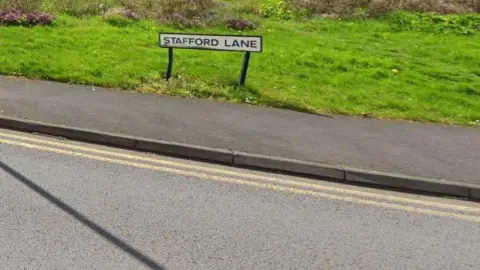 A road sign with Stafford Lane written on it stands on grass next to a pavement