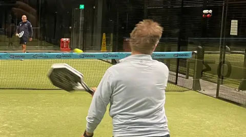 A general view of two people playing padel at an indoor court. One person, with short blond hair and wearing a grey sweater, is in action having just hit the ball. They are pictured from behind and the ball is travelling towards a man in blue shorts and sweater preparing to received the ball.