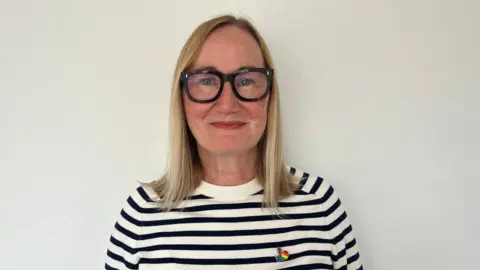 Anya Pearson A head and shoulders shot of Anya. She is wearing a black and white striped jumper and has a Pride pin on her right lapel. She has shoulder length blonde straight hair and is wearing thick framed black glasses. 