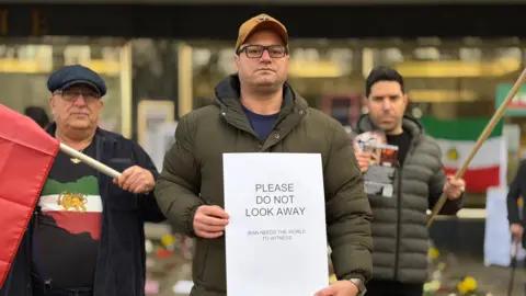 Three men are looking at the camera at the silent protest in Cheltenham. The two men in the foreground are each holding Iranian flags. The man in the front, Pasha Pour, is wearing a green puffer jacket and a brown cap. He is holding a white sheet of paper that says "please do not look away".