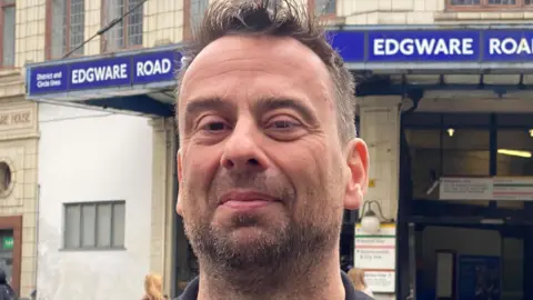 Head and shoulders image of Ben Thwaites, a man with dark brown hair and a beard. He looking into the camera. Behind him is Edgware Road Station in London. The station has a blue sign above it.