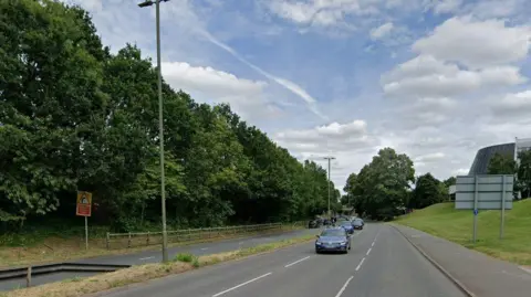 A Google Street View image showing the dual carriageway of the A25 through Guildford. There are three lanes of traffic on the near side and two on the other, with a grass verge and trees at the side of the road.