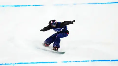 PA Media Mia Brookes wearing a Team GB snowboarding outfit, including a purple helmet, has her arms outstretched as she lands a trick on a snow slope. There are two blue lines across the white slope.