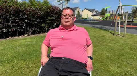 Oliver Flitcroft sitting in his wheelchair on a patch of grass with a playground behind him. Flitcroft has short dark hair and wears glasses, a pink polo shirt and black trousers.