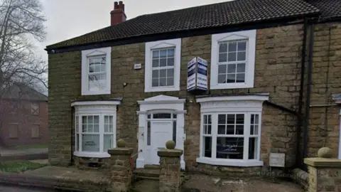 The former dental practice. It is a two-storey stone house with large windows with white panes. Dental Surgery is written on the left front bay window. There is a For Sale sign above the front door.