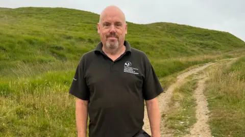 Alan Wright is standing in front of a hill footpath, he is bald and wearing a black polo top which has The Wildlife Trust logo on the front. 