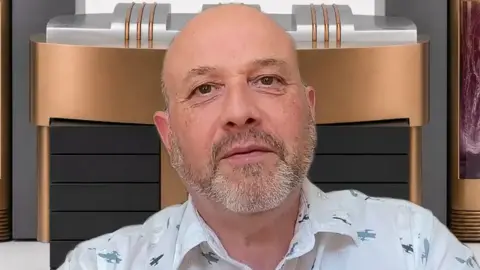 Dan Goldsmith looks at the camera - behind him is a representation of one of the supercomputers. He has a short beard and wears a white shirt with small patterns that look like spaceships.