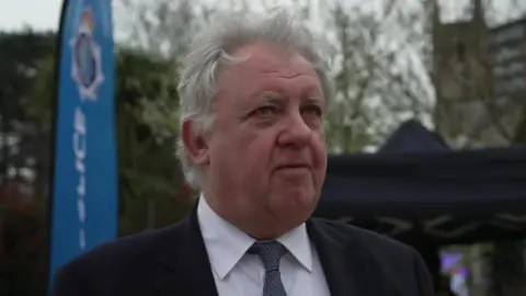 David Sidwick gives a TV interview in the street. He wears a dark suit and tie and has white, medium length hair.