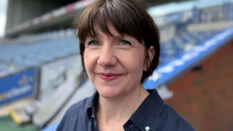 Course leader Daniella Slinger at Blackburn Rovers Ewood Park stadium with short dark brown hair wearing a navy shirt smiling.