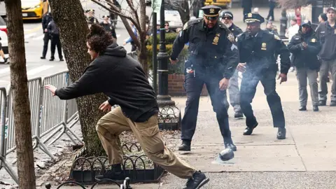 Man with curly hair wearing a black sweatshirt and beige pants runs away from police officers as smoke rises