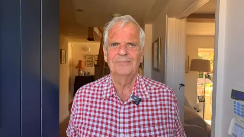 Jasmine Lowe / BBC Peter Robinson - a man in a red and white shirt. He is tanned and has white hair. He has piercing blue eyes. He is standing in the doorway of his home that looks like a cottage.