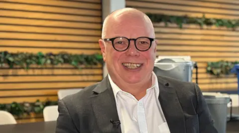 A bald man wearing black rimmed glasses, a grey suit jacket and white shirt, smiling at the camera