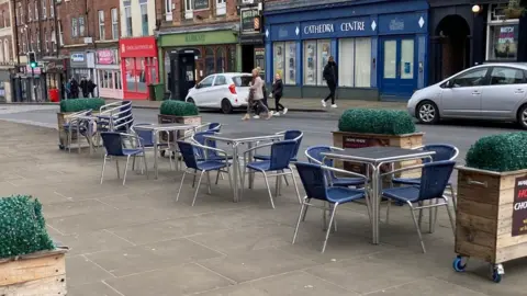 Google A city centre street with a seating area on the pavement. There are shops lining the street and people and cars pictured in the background.