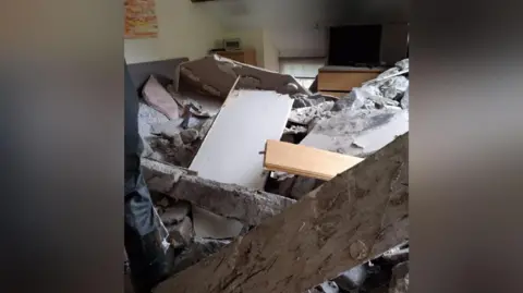 Emyr Owens A damaged bedroom with rubble covering a bed. A large fallen wooden beam can be seen in front of the bed, while two chest of drawers and a window can be seen in the background. 