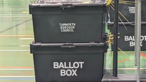 A stack of black ballot boxes marked “Tamworth Borough Council” and “Ballot Box” inside a large indoor hall with a green floor.
