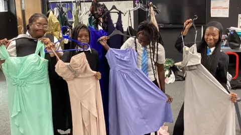 A group of young people indoors holding up long dresses on hangers, with clothing racks and costumes in the background.
