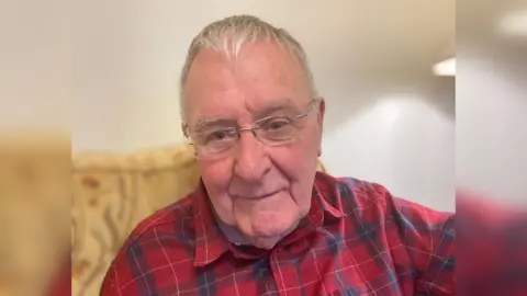A man sits on a sofa. He is wearing a red chequered shirt, eye glasses, and has grey hair.