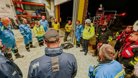 Humber Rescue Fourteen of the team stand in a circle in back at the station. The are still wearing their waterproofs and one of the life boats can be seen through a large opening in the station wall.