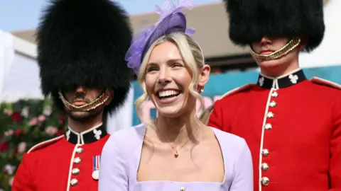 A woman in a lilac dress and fascinator smiles in front of two guardsmen wearing full parade uniform of black busby hats and red jackets with black collars