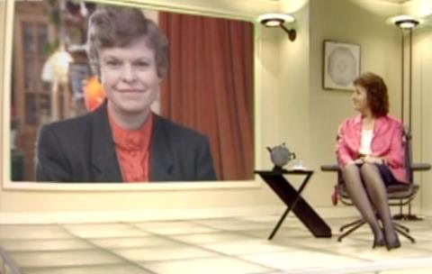 Judith Hann sitting in a chair in the set, with Christine McNulty on a large screen behind her.