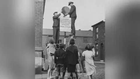 Hulton-Deutsch/Getty Images Black and white image dated 1937 of children watching two men erect a sign in a Salford street denying access to all pedal and motor vehicles to become a children's play area. 