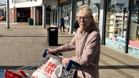 Lorraine Monaghan, a mature woman with long blonde hair with a fringe and glasses holds onto her shopping trolley outside the Braes shops. The trolley is full of bags from different shops. She wears a dusky pink jacket.