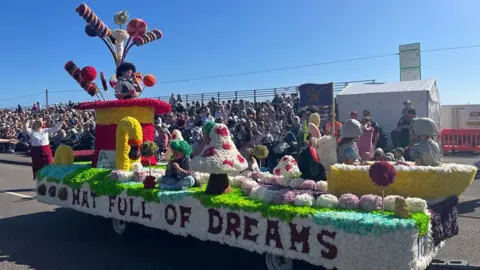 A colorful parade float decorated with large mushrooms, candy canes, and whimsical characters in costumes. The float features the text 'HAT FULL OF DREAMS' on its side. Spectators are seated in bleachers in the background, watching the parade.