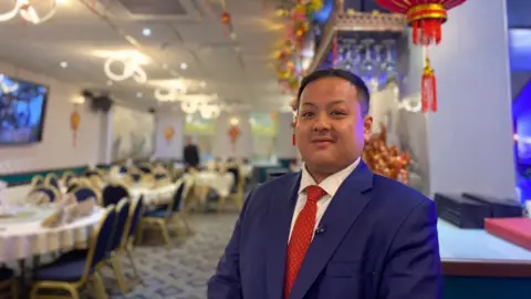 BBC / Claire Hamilton A Chinese man wears a navy blazer, red tie and white shirt, and smiles at the camera as he stands in a restaurant.