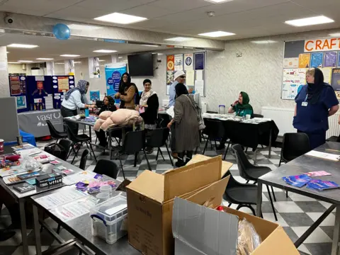 Several people in a room in the gurdwara with lots of tables and chairs.