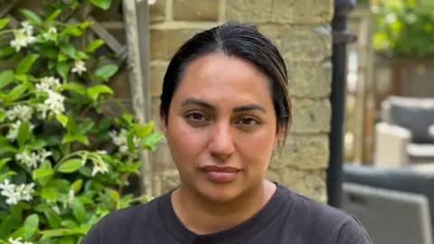 Gabby Colenso/BBC Amelia Palmer stands wearing a black T-shirt in front of brick wall and a plant with flowers to the left. To the right there is garden furniture in the distance. 