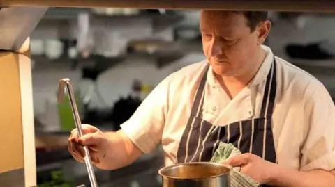 Jobe Ferguson A man in chef's outfit ladling food from a saucepan. 