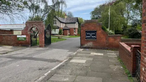 James Robinson/NCJ Media The entrance to Hirst Park which is a road with a red brick wall on either side. Black gates are open on either side of the road and there is a black sign on the right which reads 'Welcome to Hirst Park'. There is a detached house just on the inside of the gate. Trees can be seen behind the wall.