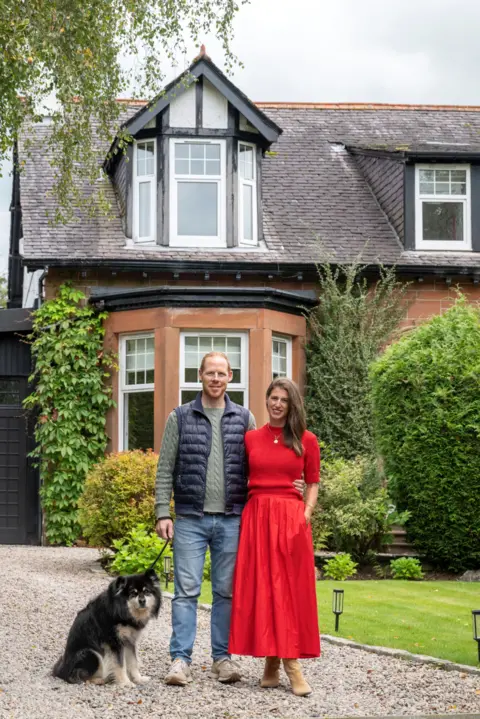 IWC Media Scott and Wendy stand side by side on a gravel driveway beside a black dog, in front of a large house with a steep, tiled roof and dormer windows.
The house features warm stone walls, black timber accents, and is surrounded by neatly maintained greenery and shrubs.
Overhanging tree branches and soft daylight create a calm, residential atmosphere in the garden setting.
