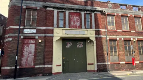 The currently derelict building of the proposed Glassworks site. It is a two-storey, red brick, old factory building with PS written on the green doors. 