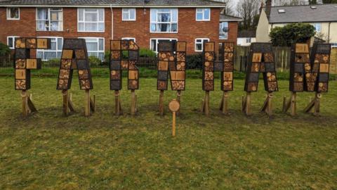 A 3D wooden sign that reads FARNHAM is stood on plinths in a meadow in Surrey, each letter is split into sections containing different habitats for insects