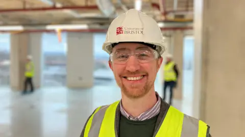 Marty Reid has a white hard hat on, with safety goggles and a hi-vis jacket. He is smiling and has a beard.